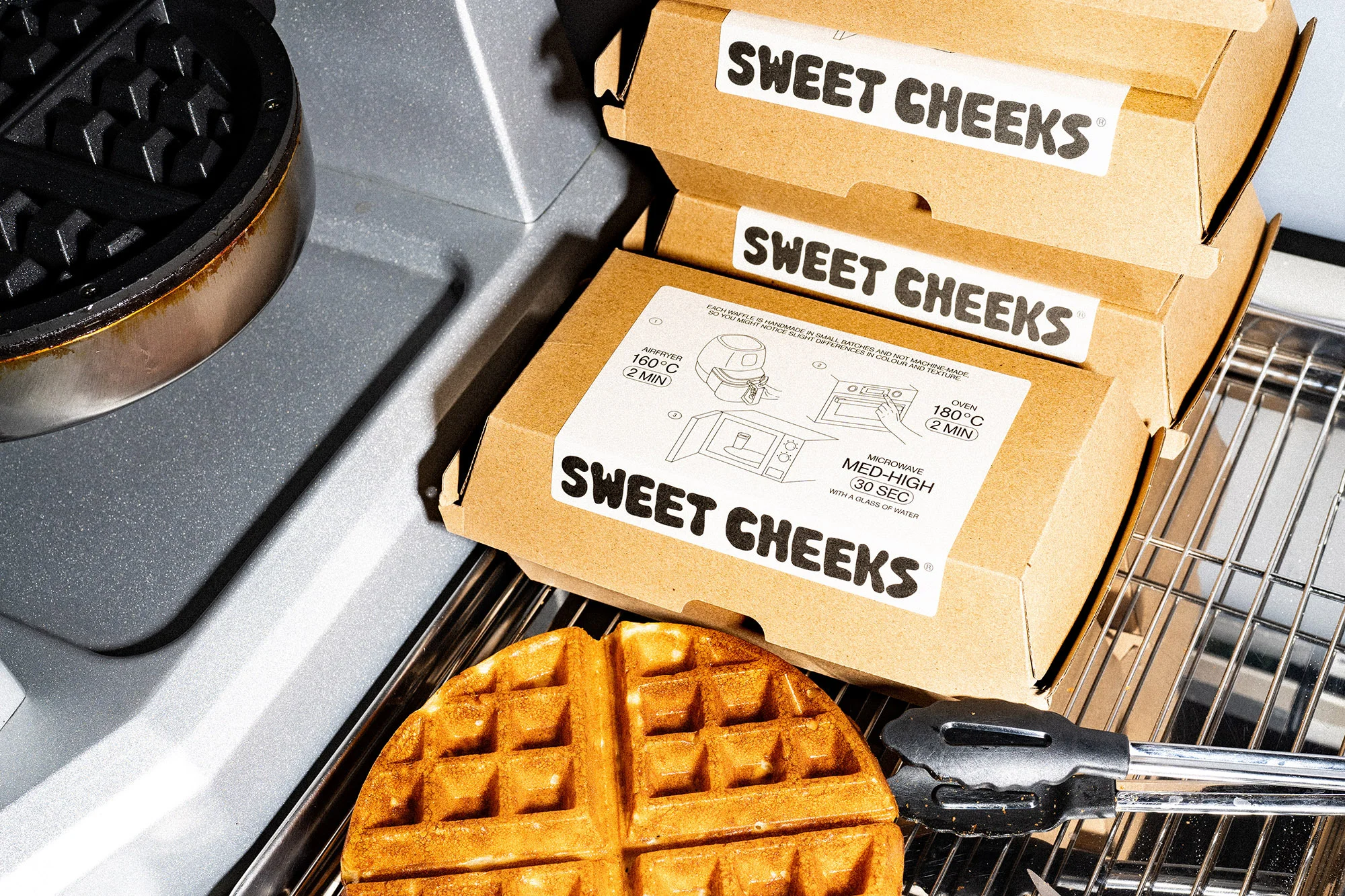 Waffle sits on a rack next to a waffle iron, with Sweet Cheeks brand boxes, tongs, and metal kitchen appliances for a food and beverage theme.