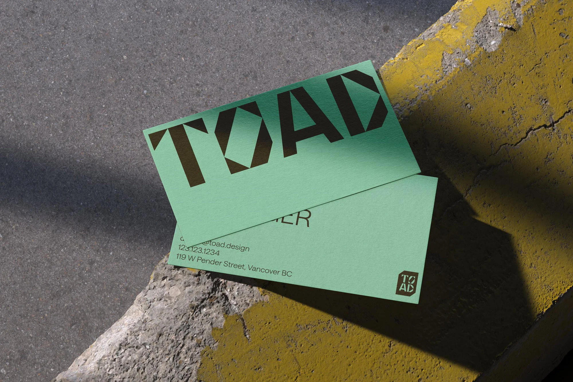 Two light green business cards with a bold sans-serif font rest on a concrete surface next to a partially painted yellow curb.