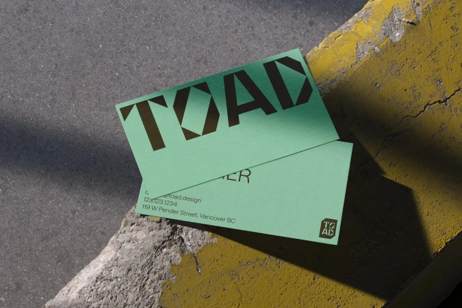 Two light green business cards with a bold sans-serif font rest on a concrete surface next to a partially painted yellow curb.