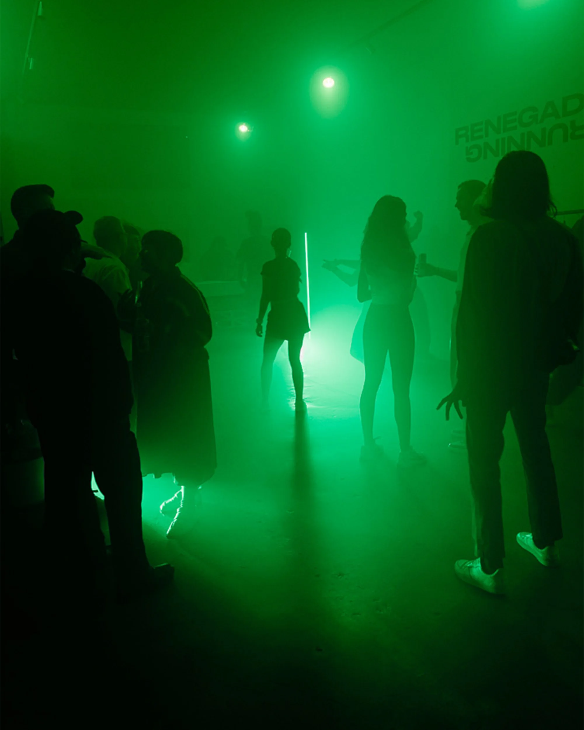 Silhouetted figures gather in a green-lit, smoky indoor venue, possibly a party or event, with blurred outlines and atmospheric lighting.