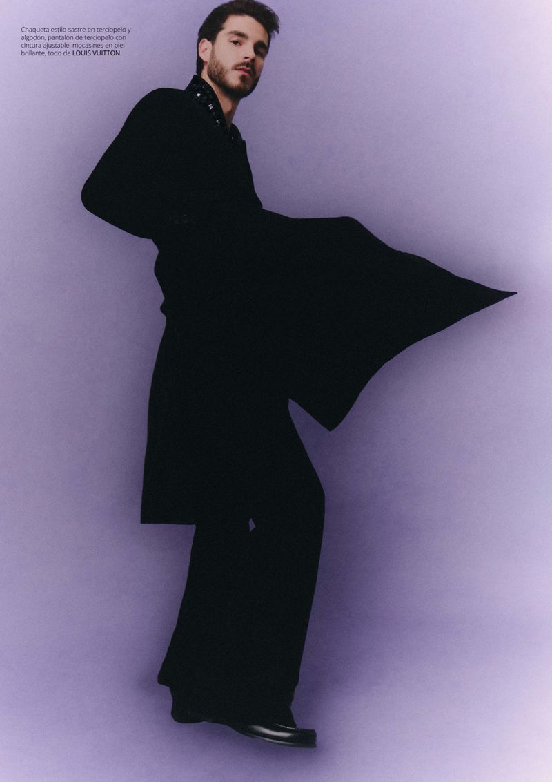 Fashion portrait of a handsome adult male model in a black coat and wide-leg trousers against a lilac backdrop in a photo studio.