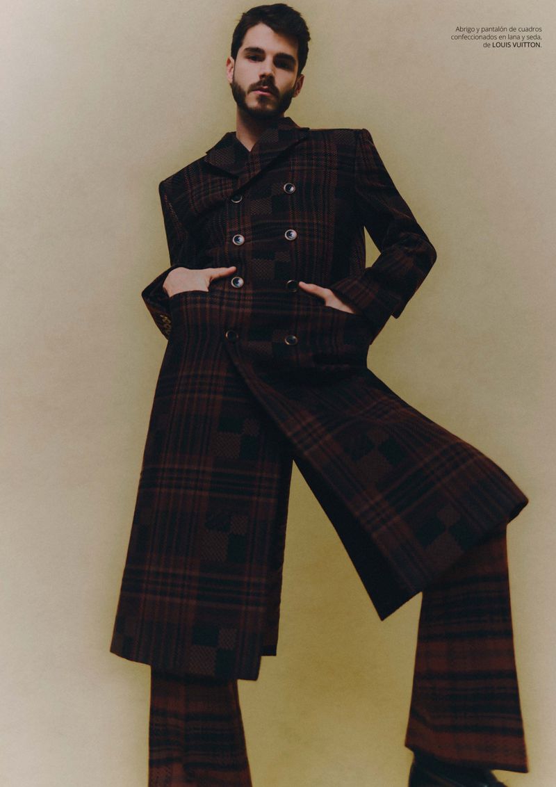 Stylish man in a brown plaid double-breasted coat and matching wide-leg pants posing against a beige backdrop in a fashion editorial shot.
