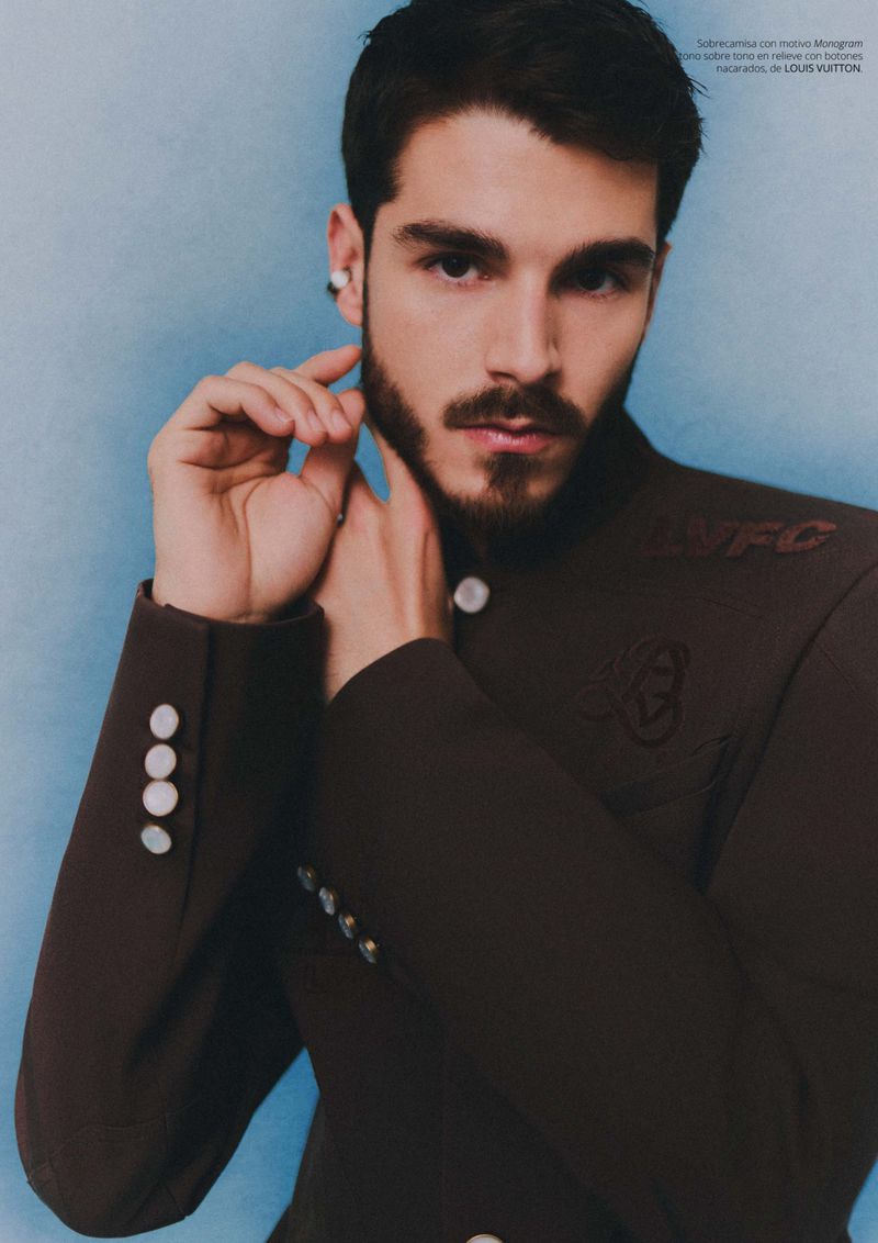 Portrait of a young adult male with a beard, wearing a brown jacket with white buttons against a blue studio backdrop.