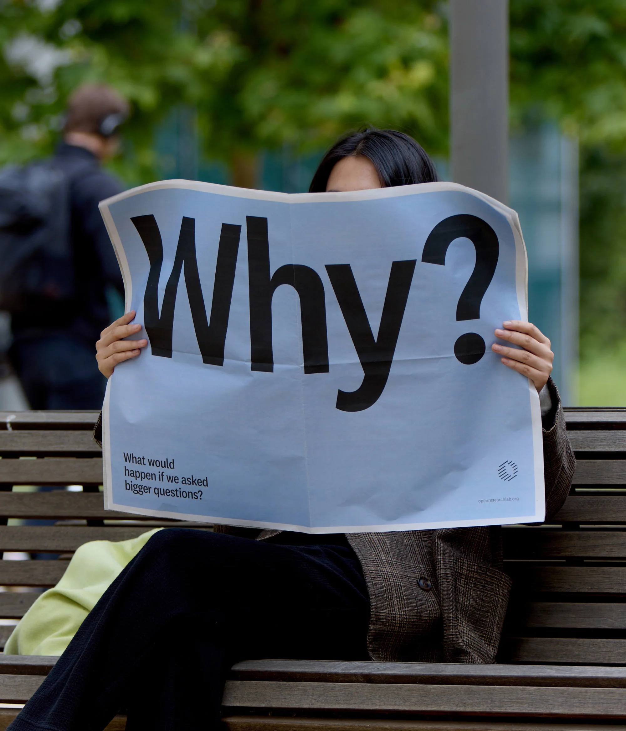 Person holding a light blue poster with bold black text reading Why in front of their face, seated on an outdoor wooden bench.