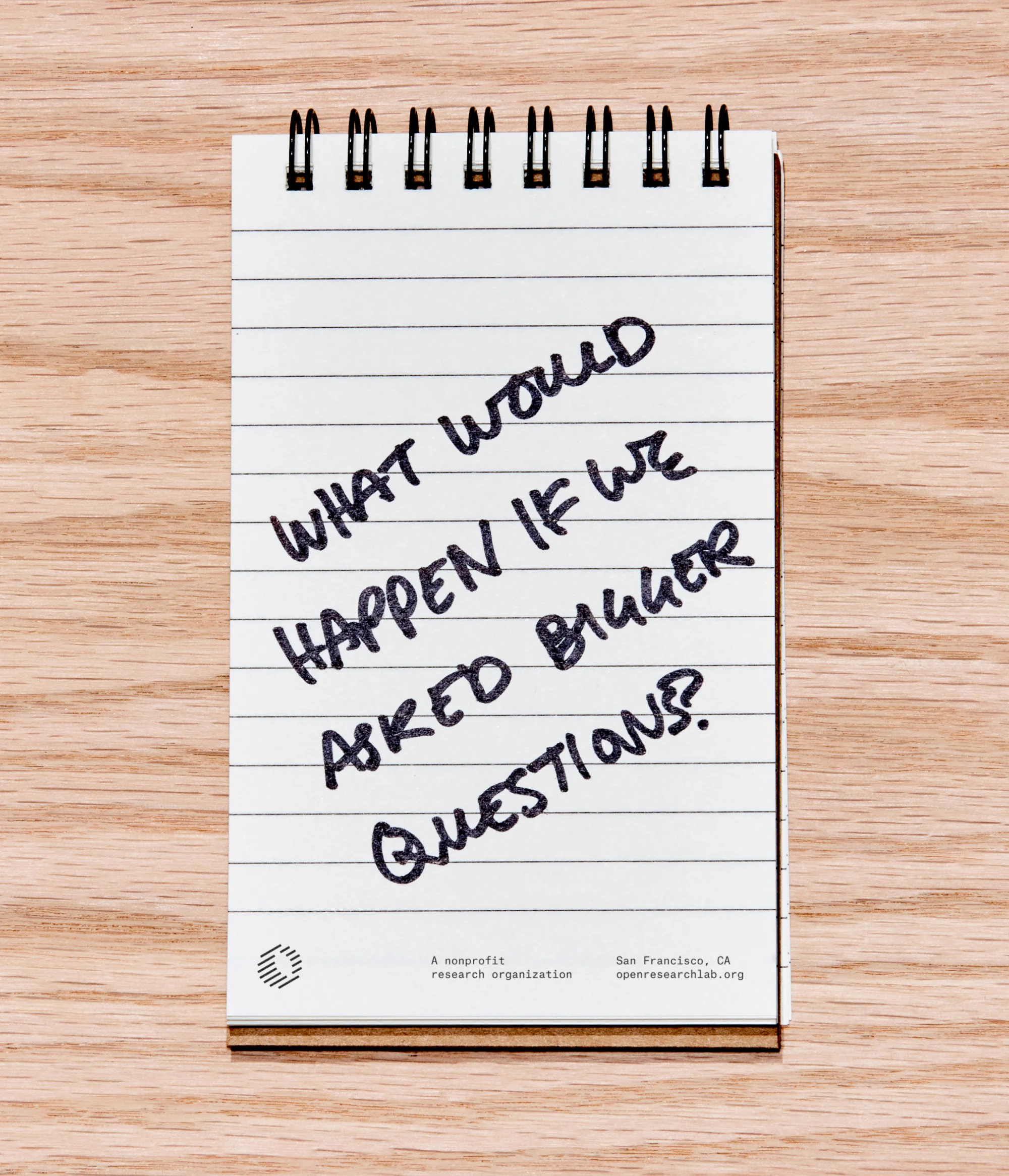 Notepad on a wood surface features the handwritten phrase, "What would happen if we asked bigger questions?" in dark ink.