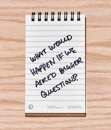 Notepad on a wood surface features the handwritten phrase, "What would happen if we asked bigger questions?" in dark ink.