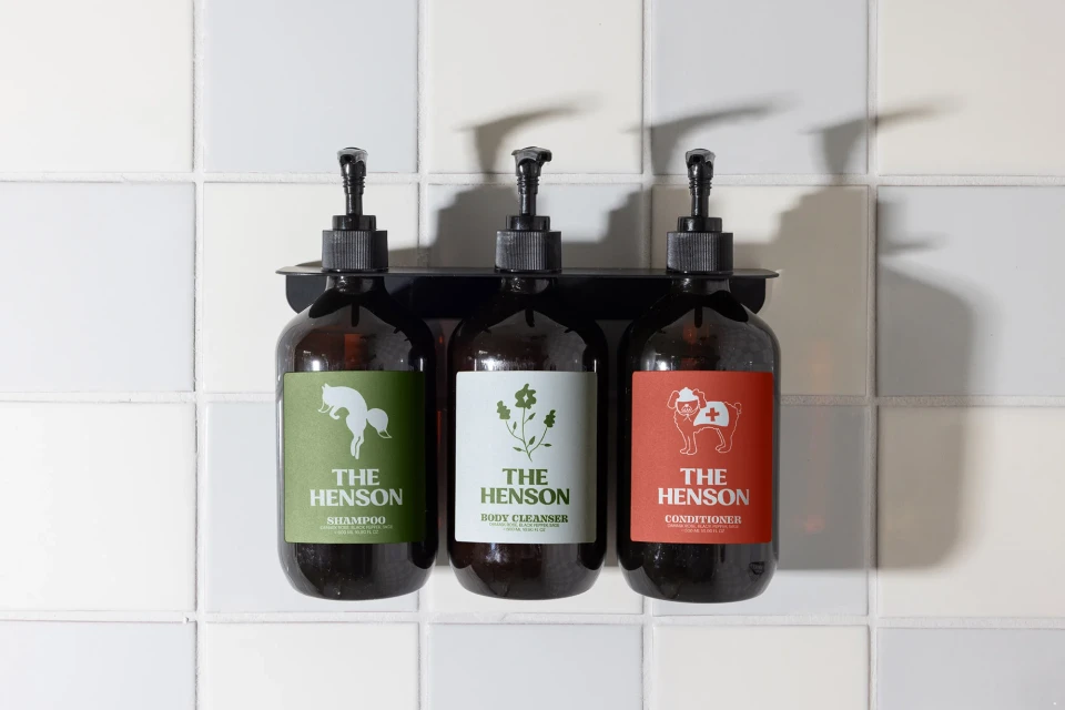 Three amber glass pump bottles labeled 'The Henson' containing shampoo, body cleanser, and conditioner against a tiled wall.