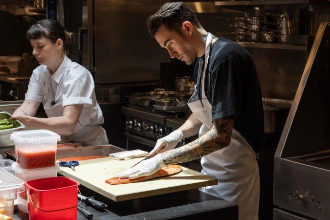 Two chefs in a commercial kitchen prepare food. One chef cuts salmon on a wooden board, while another sorts produce. Stainless steel equipment.