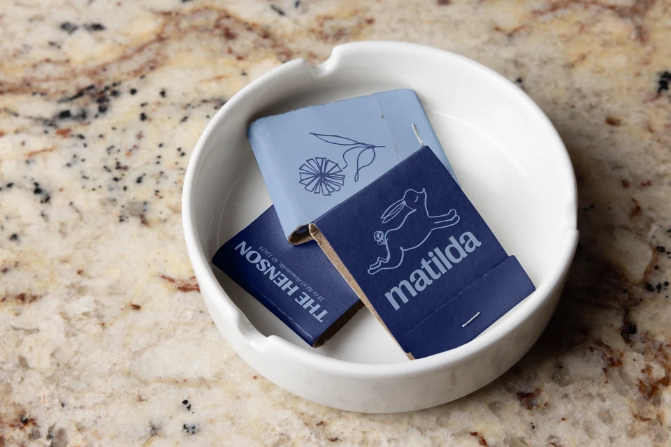 White ceramic ashtray containing matchbooks with graphic prints on a speckled tan and brown countertop surface.