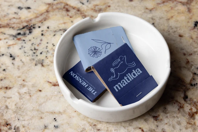 White ceramic ashtray containing matchbooks with graphic prints on a speckled tan and brown countertop surface.