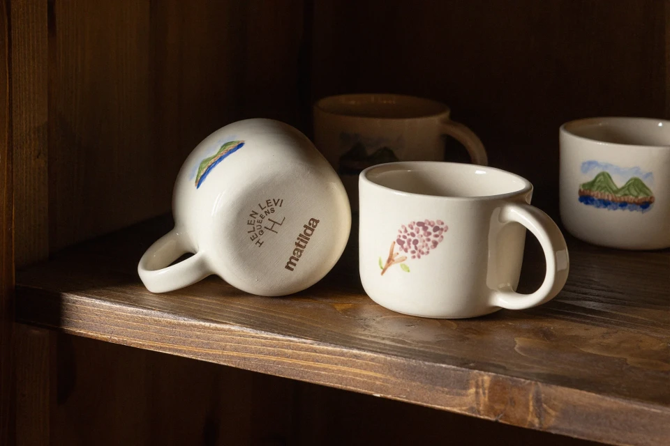 Close-up of four white ceramic mugs with hand-painted floral and landscape designs arranged on a rustic wooden shelf inside a cabinet.
