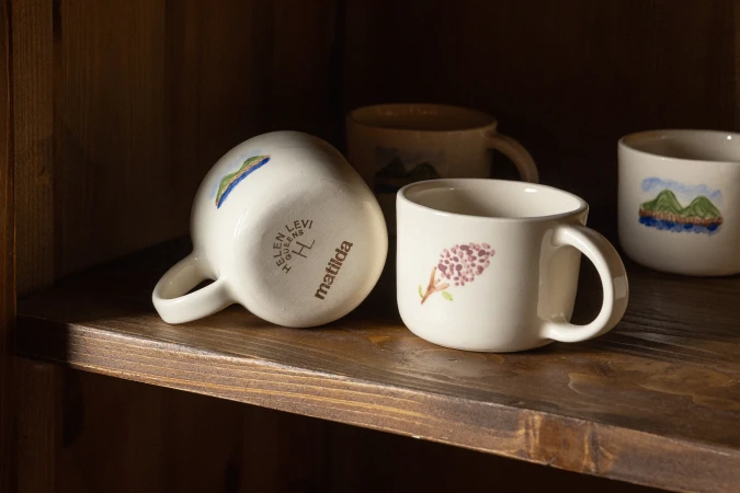 Close-up of four white ceramic mugs with hand-painted floral and landscape designs arranged on a rustic wooden shelf inside a cabinet.