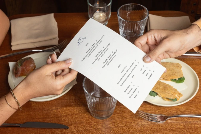 Close-up of a restaurant table with two people holding a menu, featuring dishes like roasted potato mousse, sweet milk ice cream, and savory sandwiches.