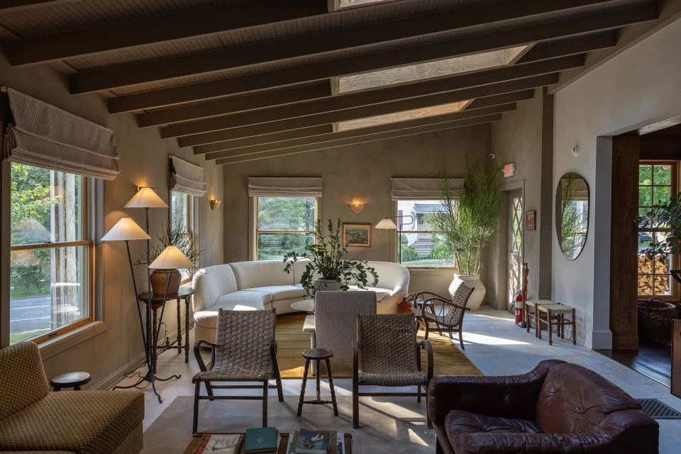 Indoor lounge area featuring a white sectional sofa, woven chairs, lamps, and plants, bathed in natural light from large windows.
