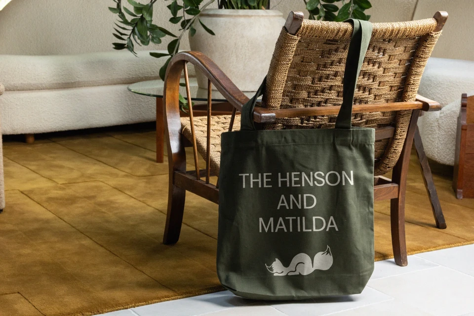 Olive green tote bag with white text hangs on a wood chair in a brightly lit, modern interior with beige walls and carpet.
