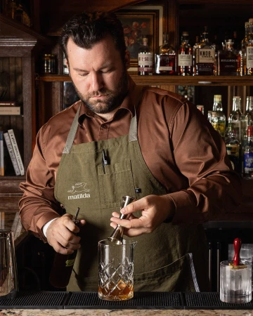 Bartender in a warm-toned bar mixes a cocktail with a jigger and mixing glass, bottles in background, creating an inviting atmosphere.