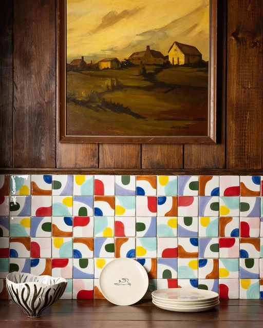 Interior shot of a wooden paneled wall with colorful tiles, a landscape painting, plates, and a decorative bowl on a shelf.