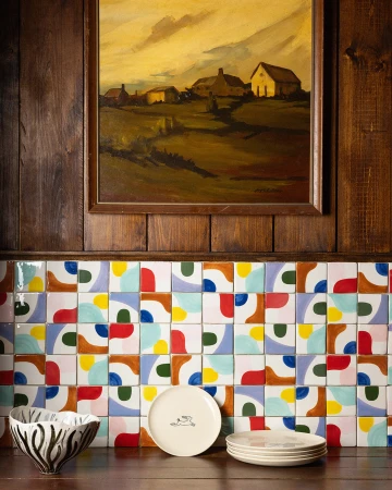Interior shot of a wooden paneled wall with colorful tiles, a landscape painting, plates, and a decorative bowl on a shelf.