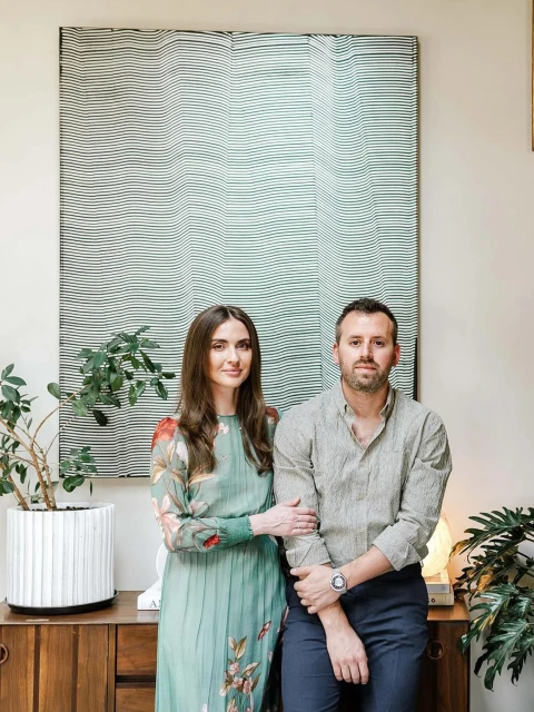 A stylish couple poses indoors next to a credenza with a potted plant and lamp, with a large abstract art piece hanging above.