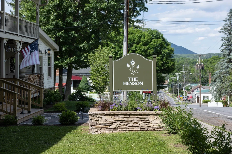 Exterior view of The Henson sign with stone base and flowers, an American flag waving on nearby building, street in the distance.