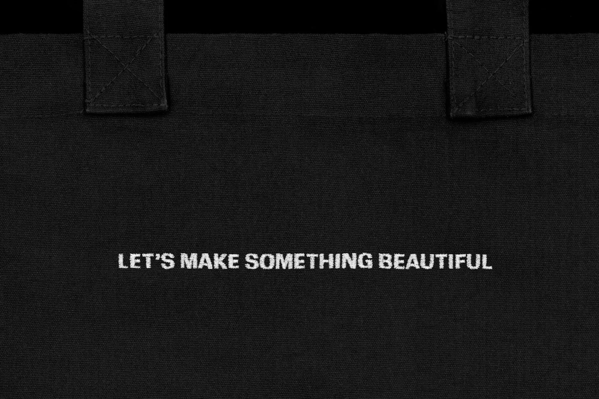 Close-up of a black canvas tote bag featuring white embroidered text 'LET'S MAKE SOMETHING BEAUTIFUL' with handles at the top.