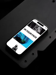 A smartphone displaying a web layout titled IN YOUR FACE rests on a dark, textured industrial surface.