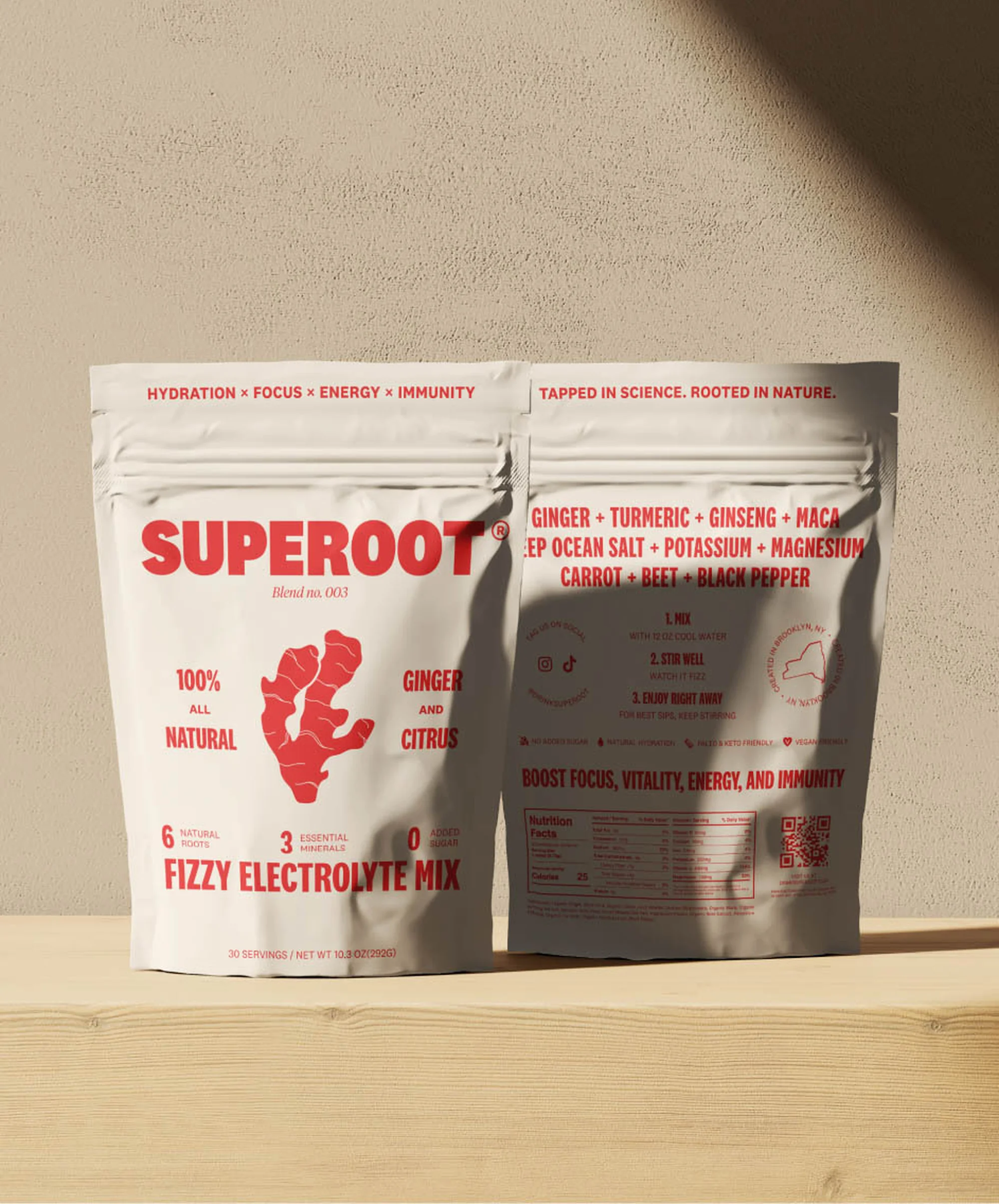 Two white pouches of Superoot fizzy electrolyte mix sit on a wooden surface against a neutral background, showcasing the product's branding and ingredients.