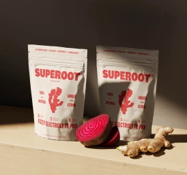 Two Superoot electrolyte pouches standing on a wooden surface beside a sliced beetroot and ginger root