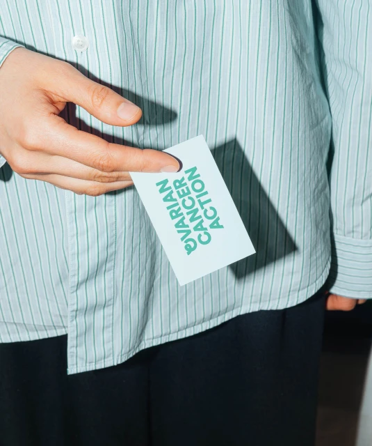Hand holds Ovarian Cancer Action card, showing support and awareness in close-up shot indoors with a striped shirt.