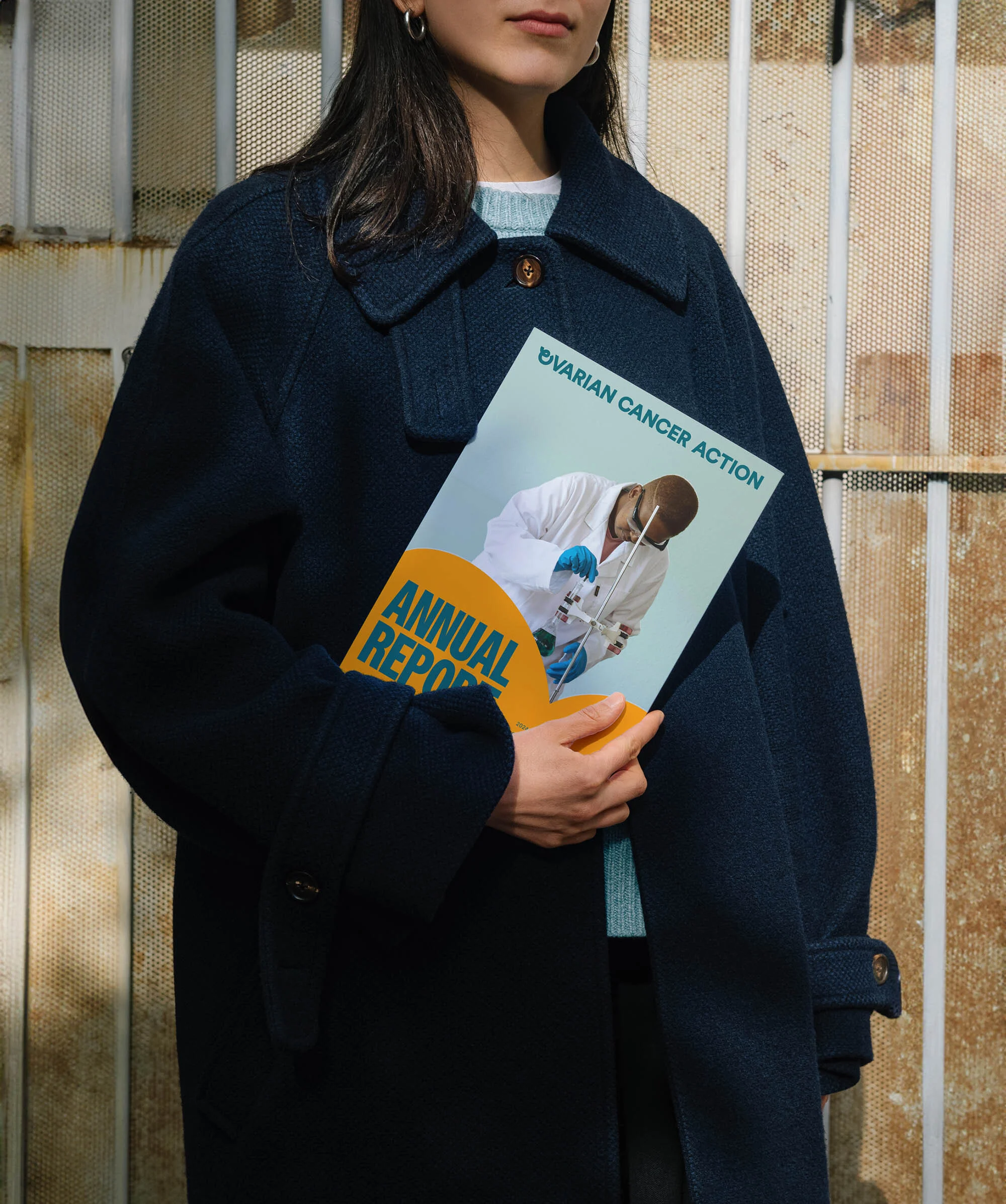 A woman in a blue coat stands against a textured wall, holding an Ovarian Cancer Action annual report, in a medium shot.