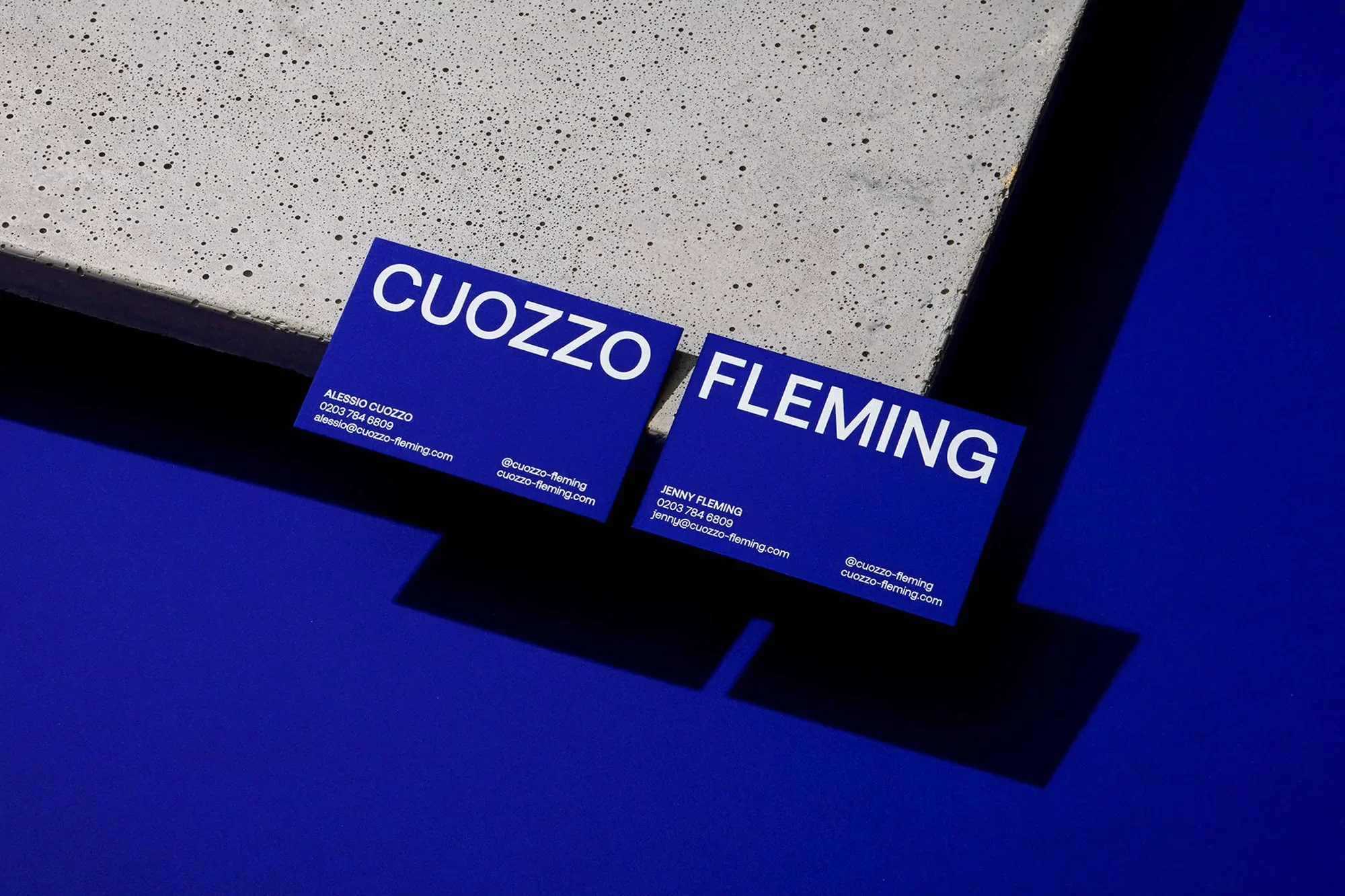 Two blue business cards with white text: CUOZZO and FLEMING, displayed on a concrete surface against a solid blue background.