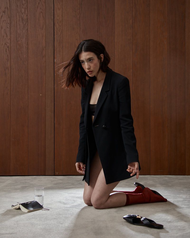 Woman in bra and blazer kneeling on the floor in front of wood paneled wall with notebook, glass of water, and red ankle boots.