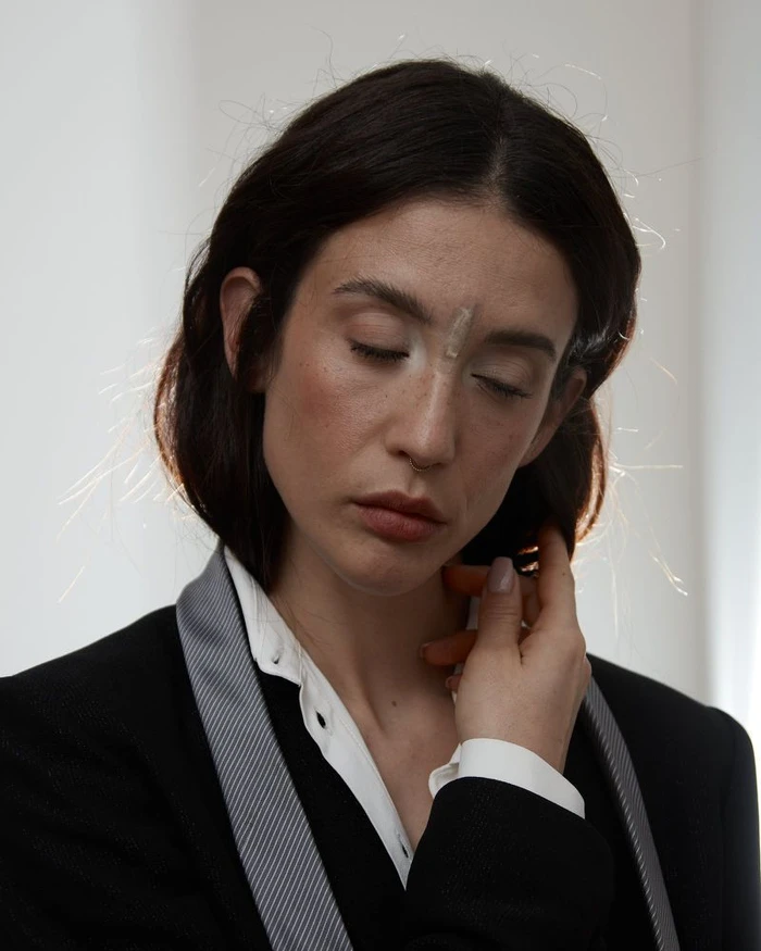 Portrait of Maria Pedraza with dark hair touching her collar, wearing a black blazer and white shirt against a neutral background.