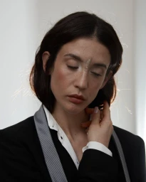 Portrait of Maria Pedraza with dark hair touching her collar, wearing a black blazer and white shirt against a neutral background.
