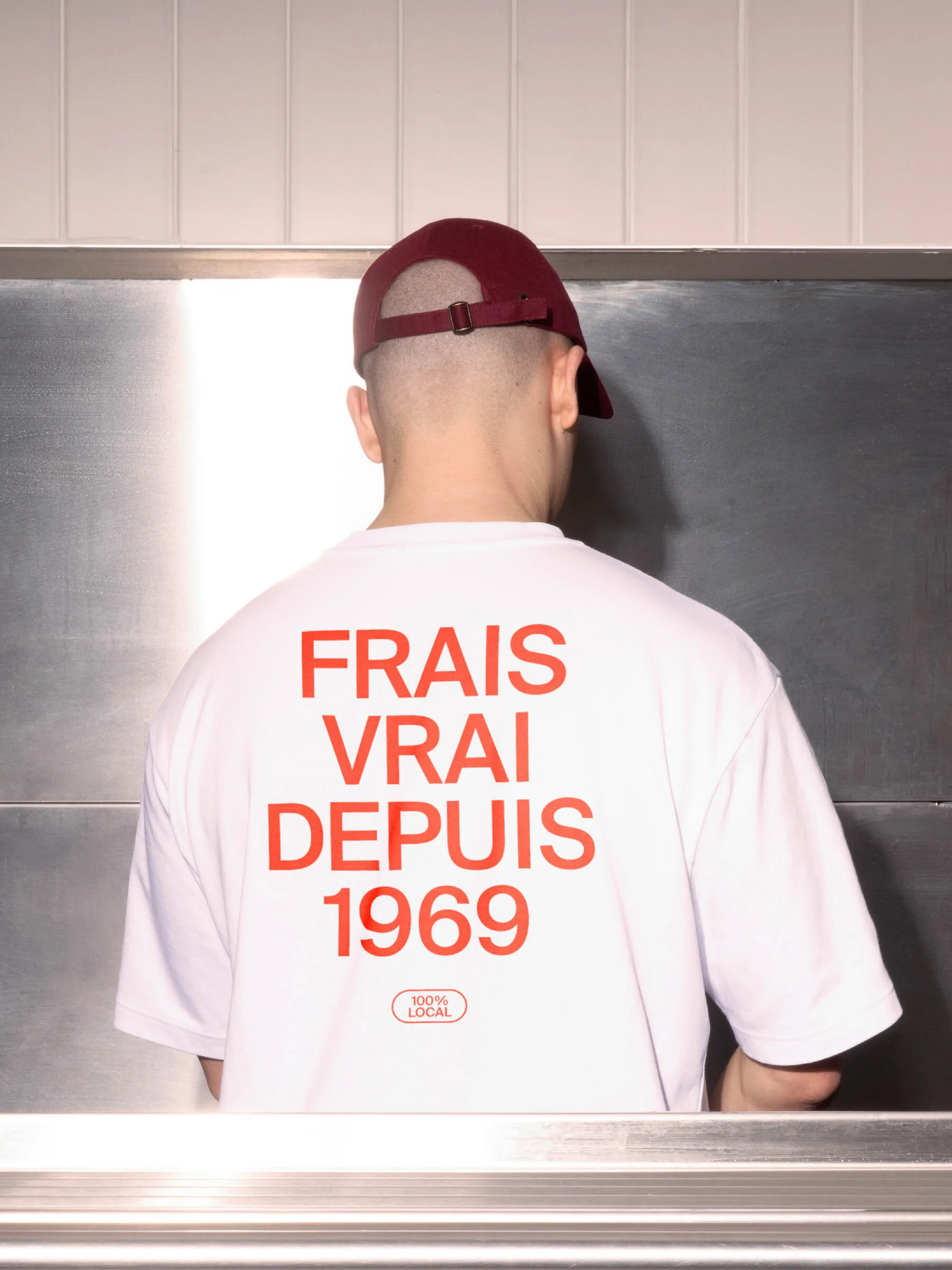 Man with shaved head wears maroon cap and white t-shirt with FRAIS VRAI DEPUIS 1969 in red lettering against stainless steel background.