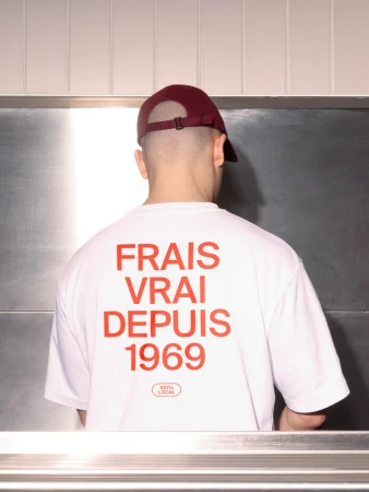 Man with shaved head wears maroon cap and white t-shirt with FRAIS VRAI DEPUIS 1969 in red lettering against stainless steel background.