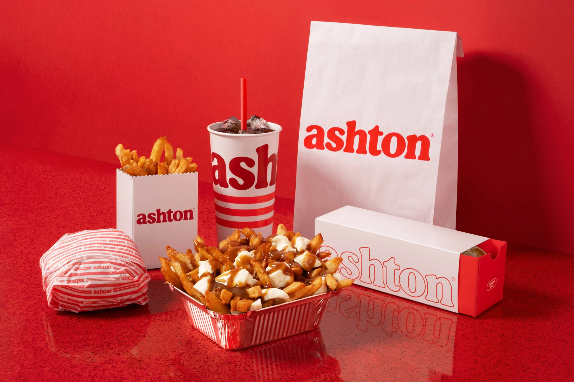 Still life of fast food items including a burger, fries, poutine, and a drink, all branded with the 'Ashton' logo on a red surface.