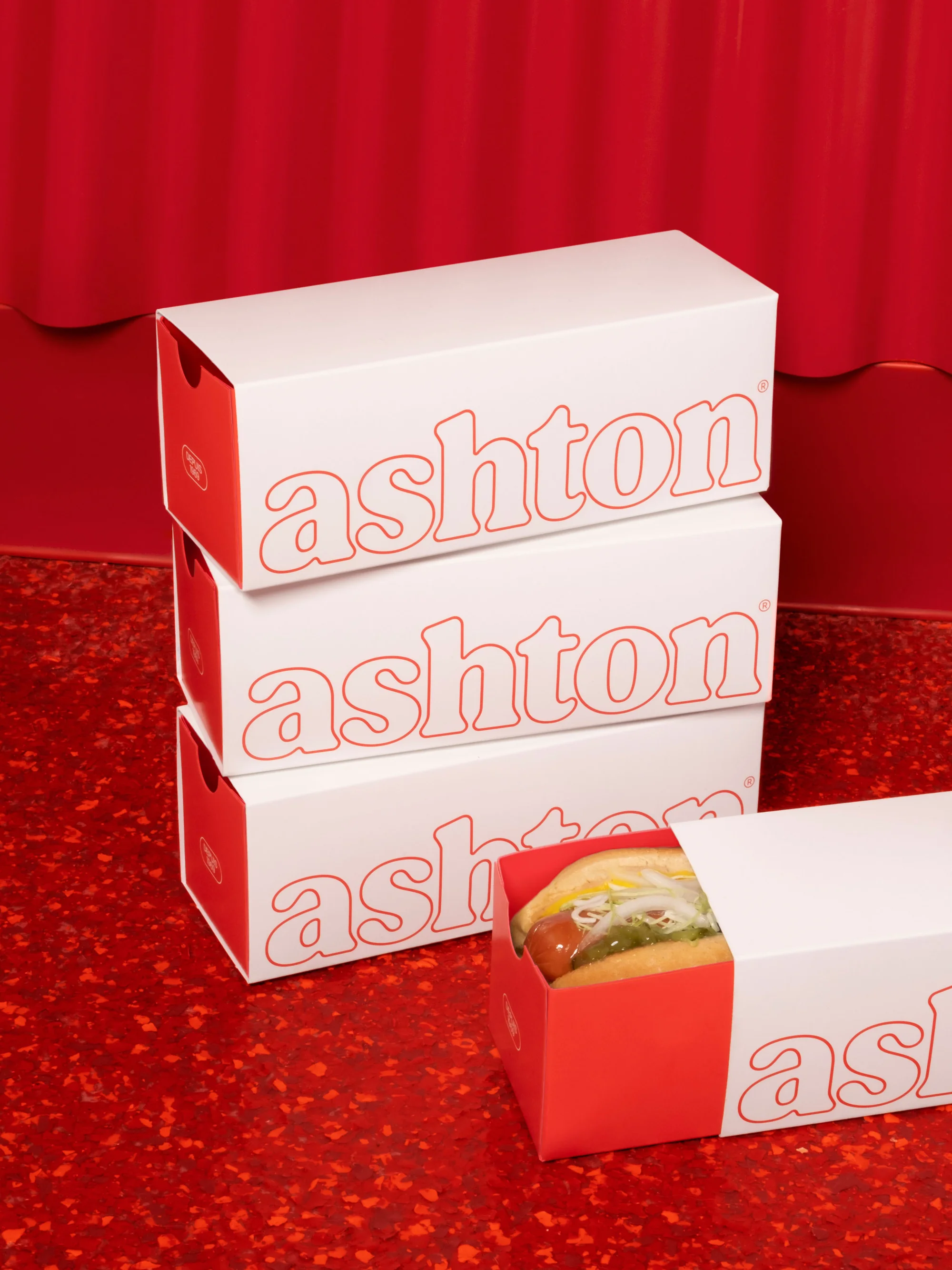 A stack of white and red branded food boxes on a red surface, with a hot dog visible in one box, under bright lighting.