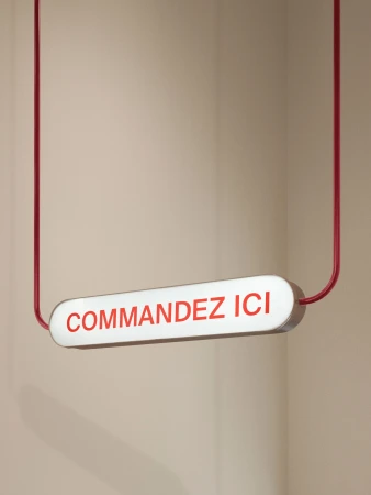 Illuminated sign suspended by red cables against a beige wall, displaying the French text "COMMANDEZ ICI" in red letters.