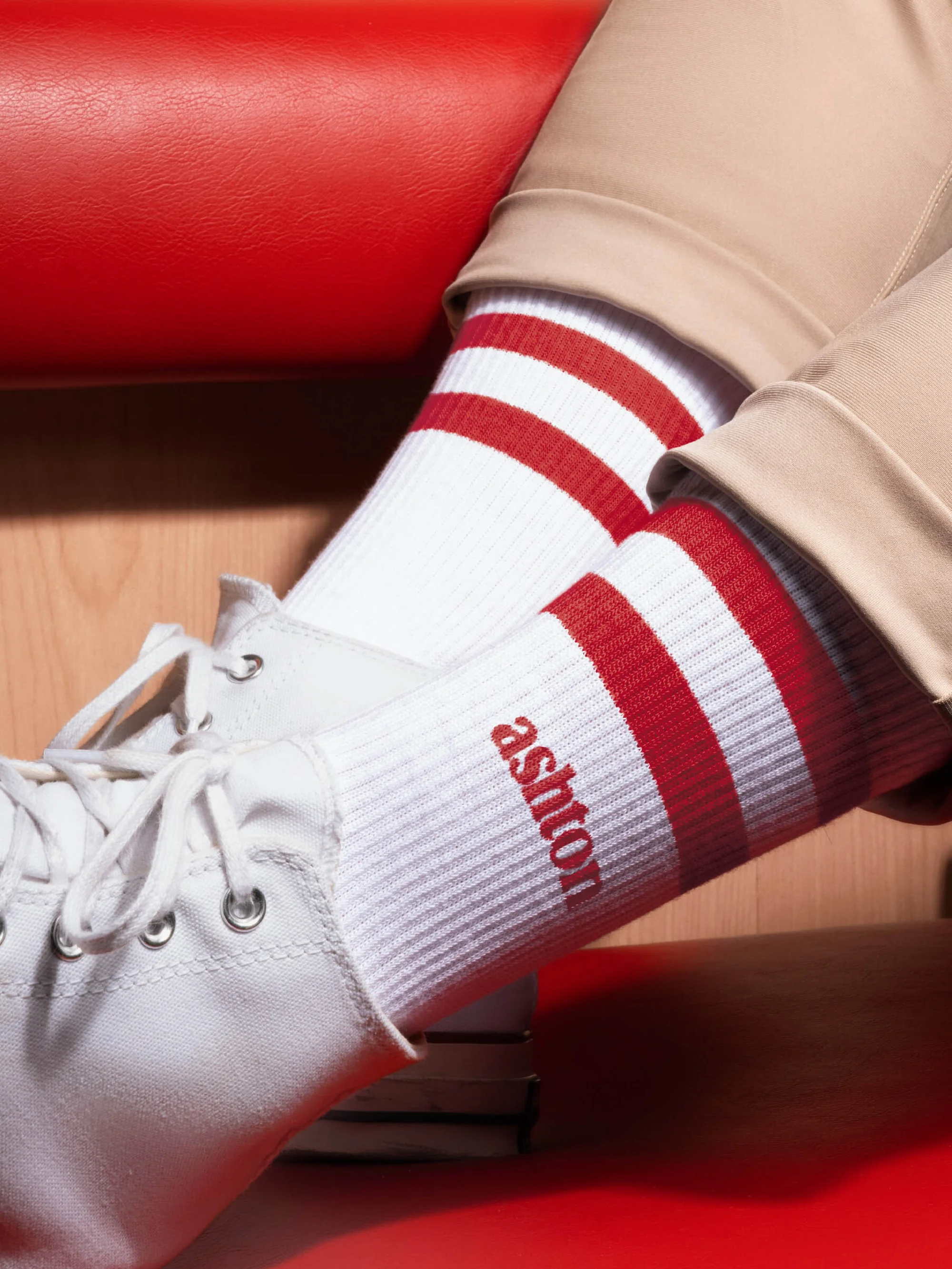 Close-up of white ribbed socks with red stripes and the name 'ashton' in red lettering, paired with white canvas shoes.