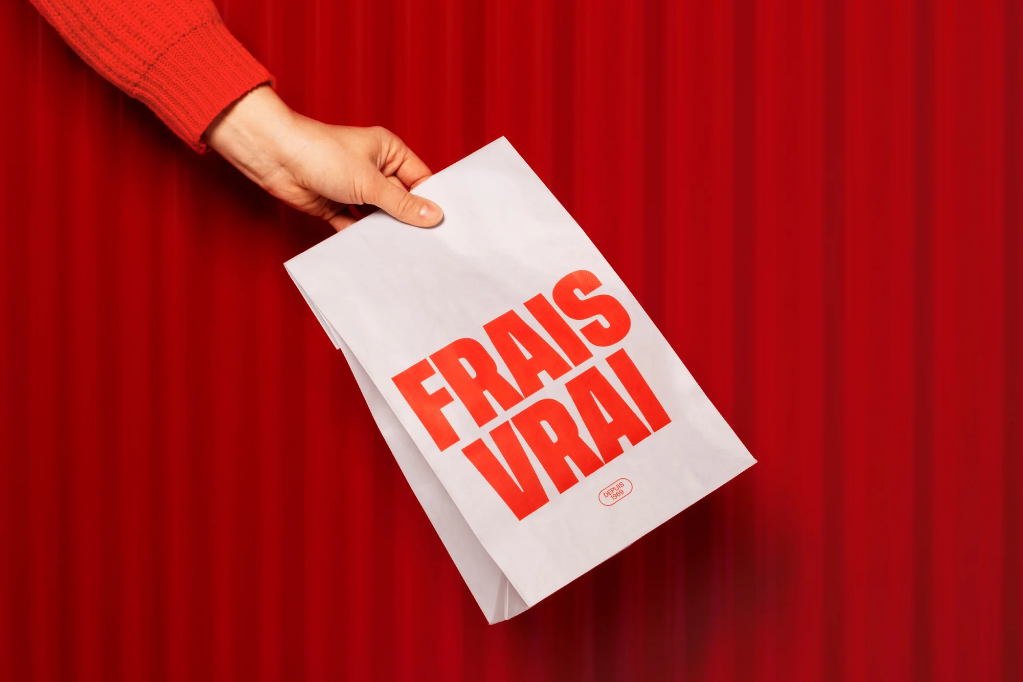 An adult hand presents a white paper bag with the French text 'Frais Vrai' in red, against a red curtain backdrop.