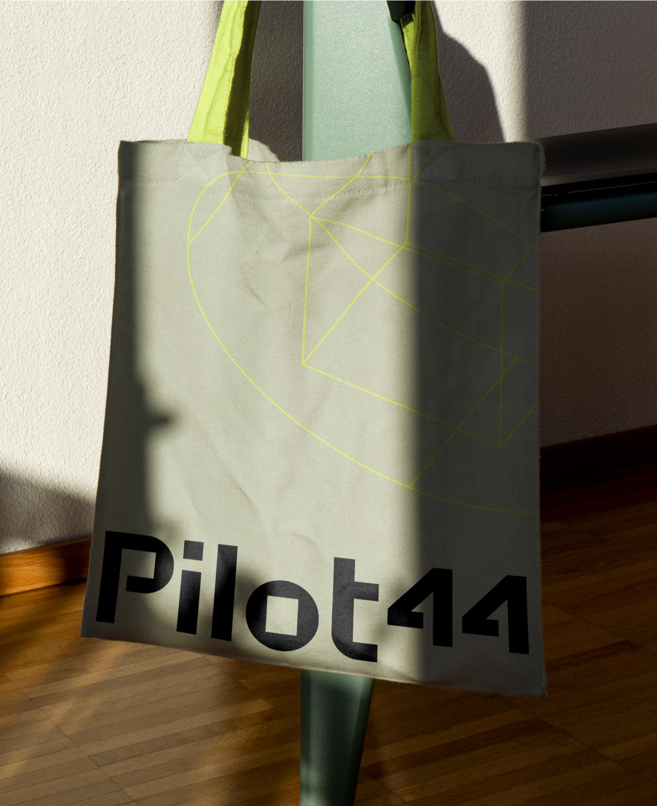 Khaki-colored tote bag with a neon green handle and geometric design. The bag features the black text 'Pilot44' on its lower half.