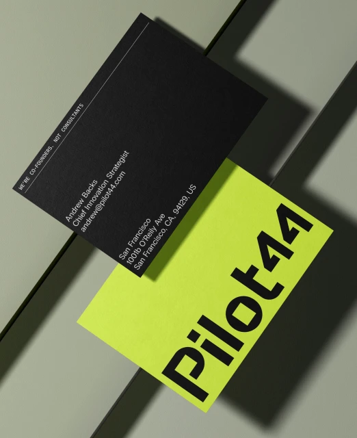 Black and neon yellow business cards stacked on a grey surface. Text includes name, address, title, and company name Pilot44.