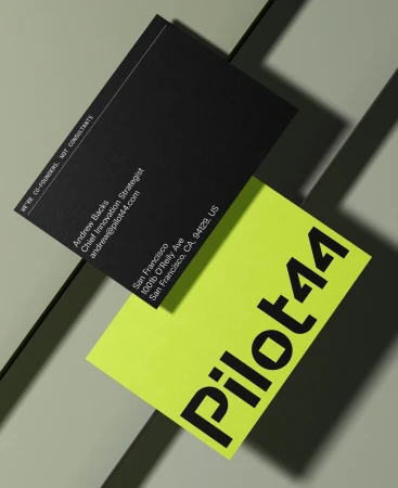 Black and neon yellow business cards stacked on a grey surface. Text includes name, address, title, and company name Pilot44.