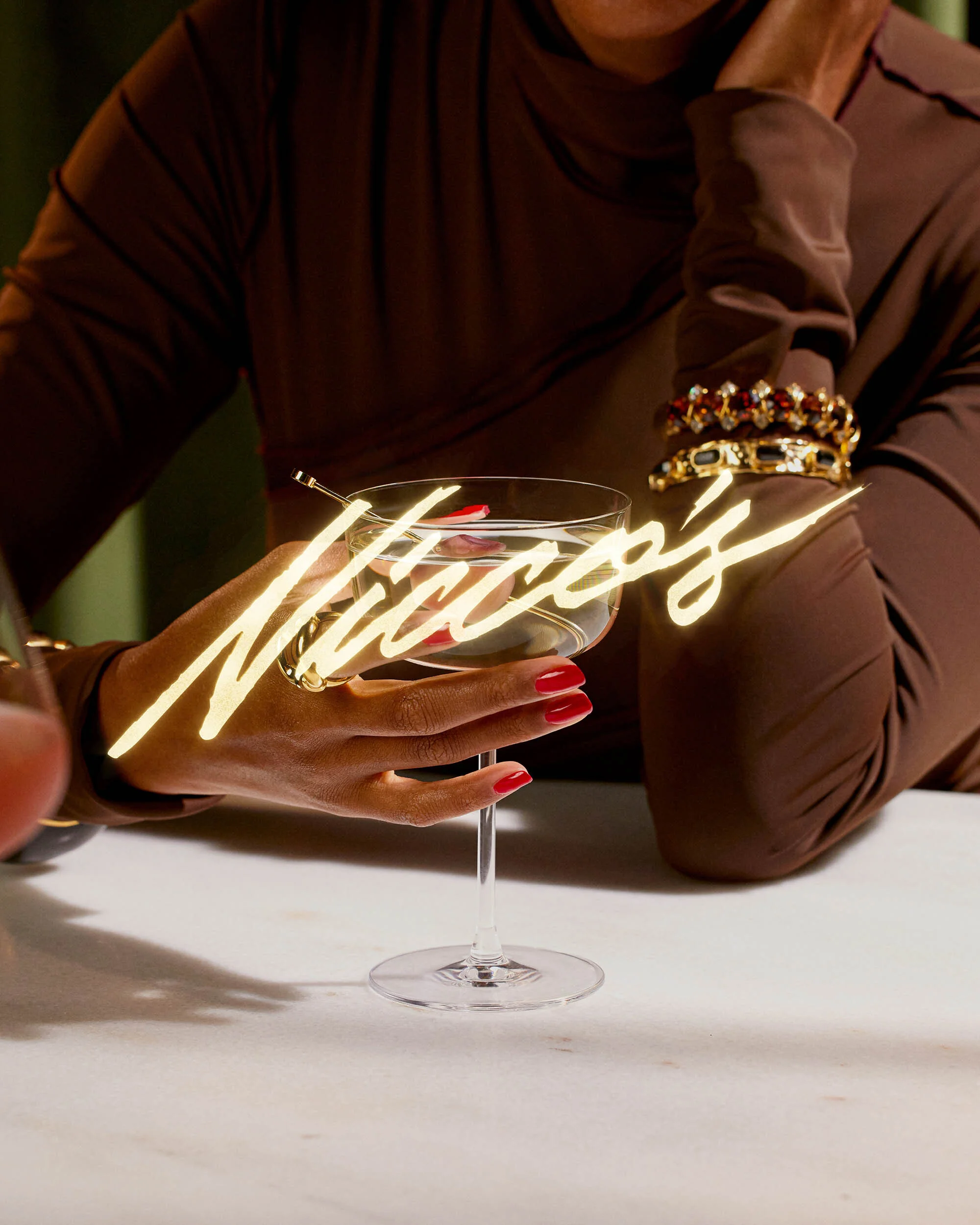 Woman with brown dress holds a cocktail glass with red nails on a white marble table, featuring jewelry and stylish presentation.