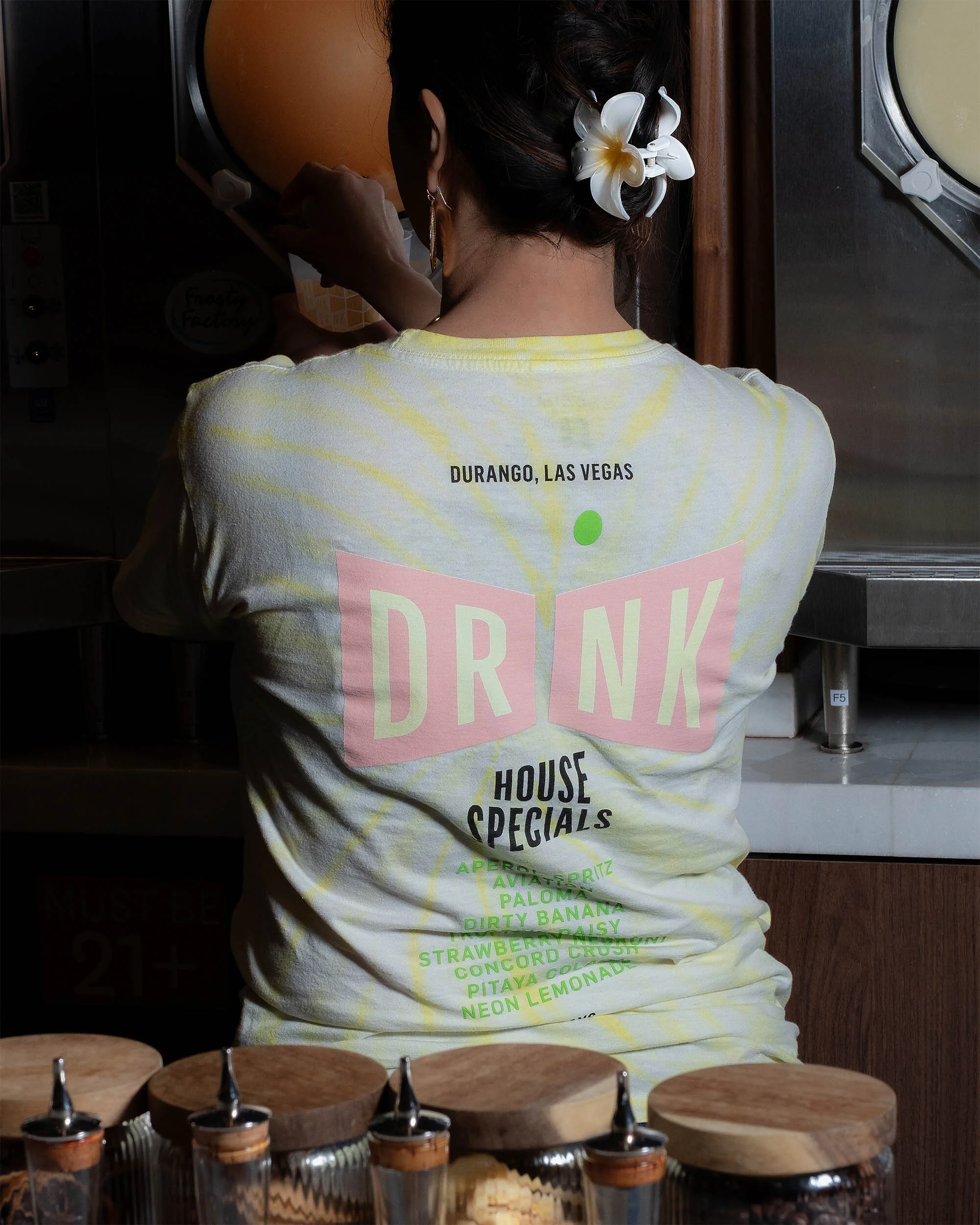 Back view of a woman in a printed t-shirt with a flower in her hair, next to a drink machine and liquor ingredient containers.