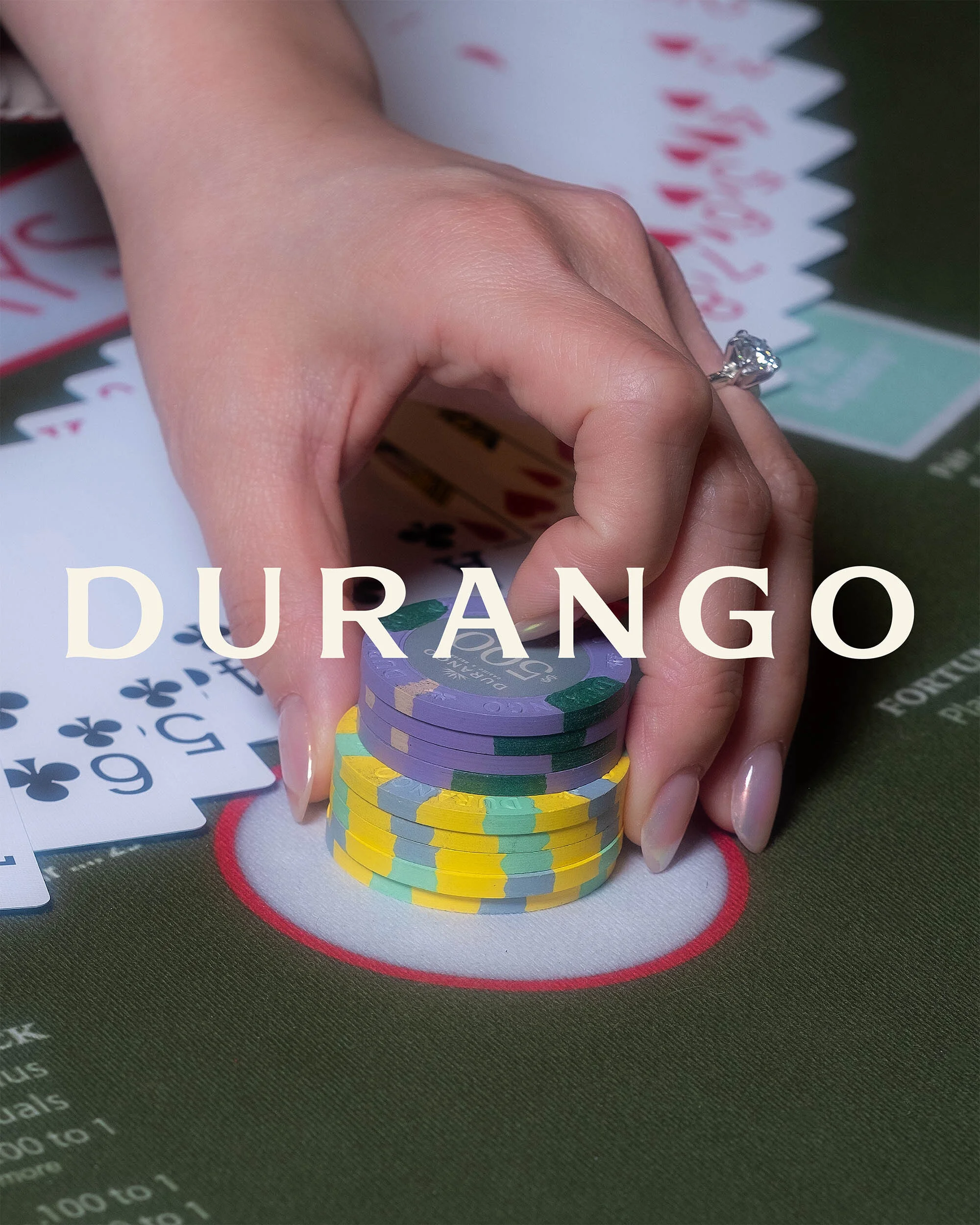 Close-up of a woman's hand with a diamond ring holding casino chips on a green gaming table with scattered playing cards.