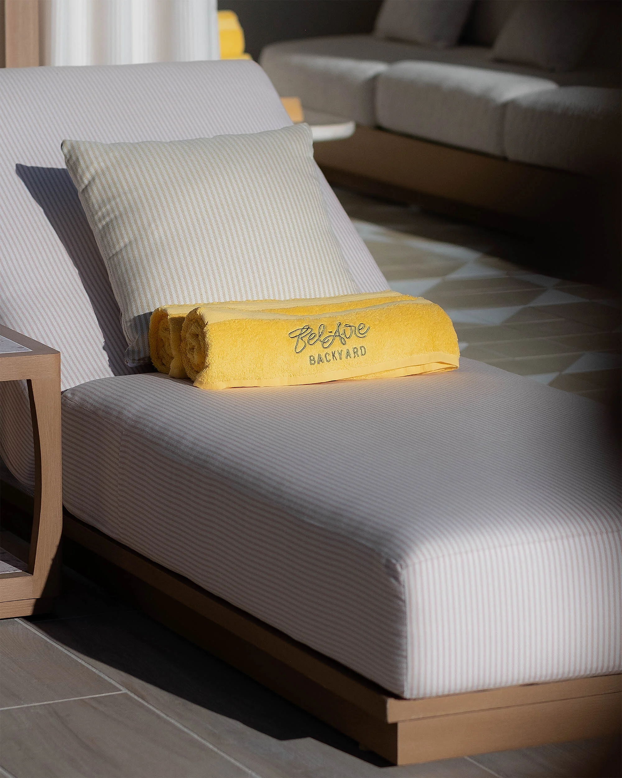 Lounger with cushion and yellow towel featuring Bel-Aire Backyard branding, set indoors with natural light.