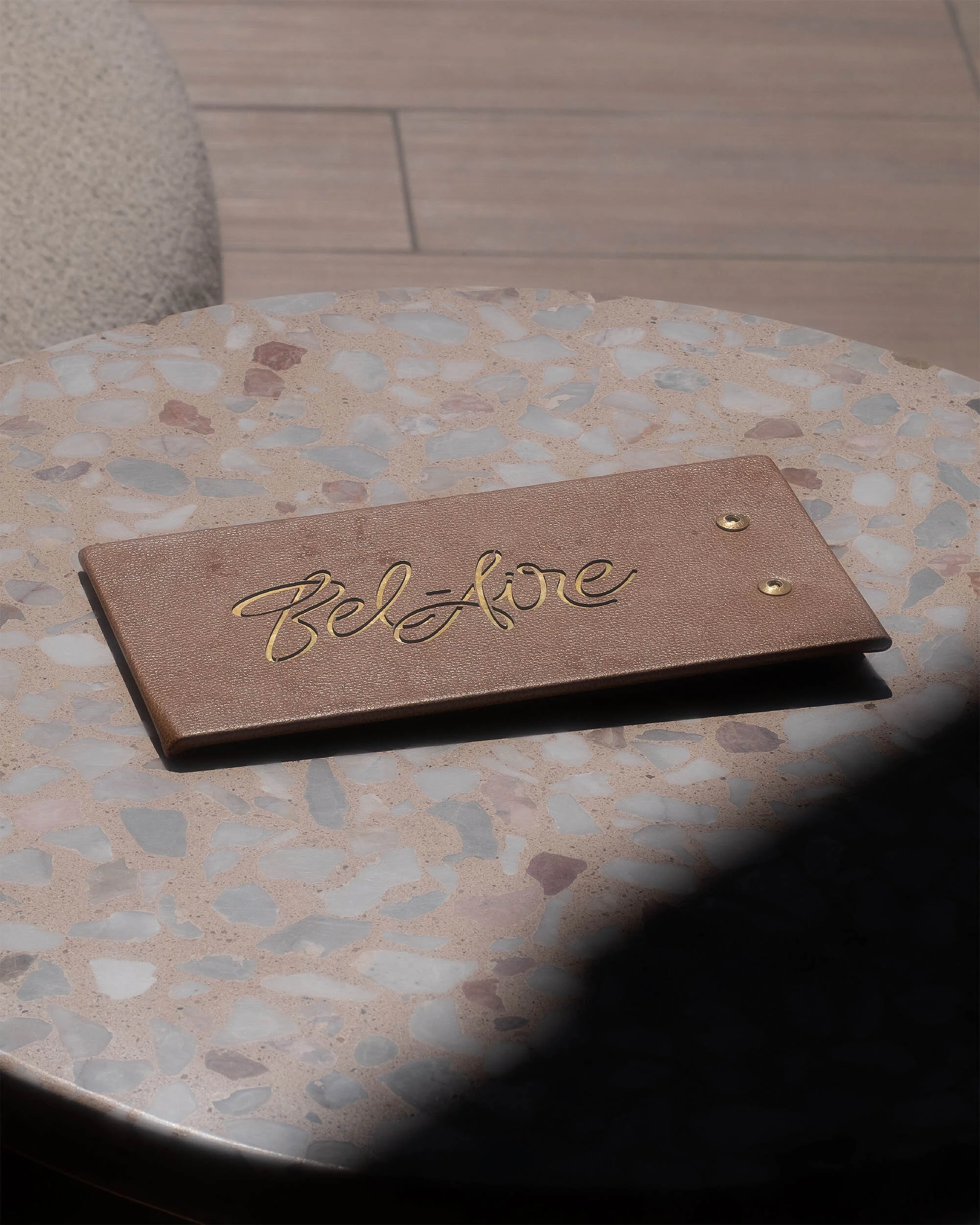 Close-up of a tan rectangular sign with the word 'Bel-Aire' in gold script, displayed on a speckled terrazzo table.