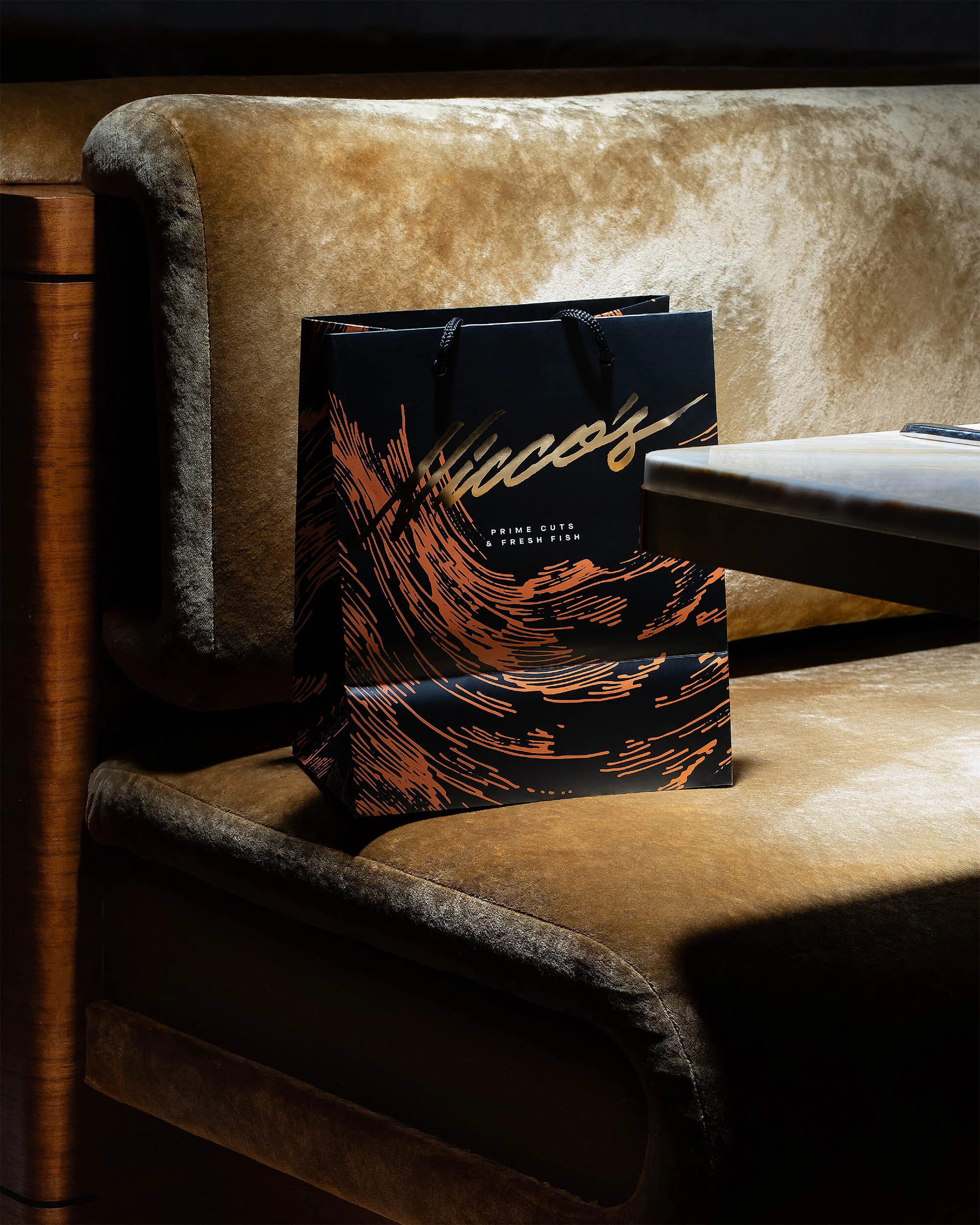Black and copper paper shopping bag on a plush tan bench seat next to a marble table in warm indoor lighting.